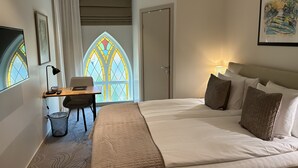 A hotel room with a large bed, a desk with a chair, a small trash bin, a stained glass window, and a door. - Sockerslottet hotell (Kristinehamn)