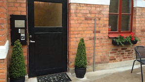 A black door with a glass panel, a keypad, and a small sign. There are two potted plants on either side of the door, a window with a red frame, and a chair outside. - Sockerslottet hotell (Kristinehamn)