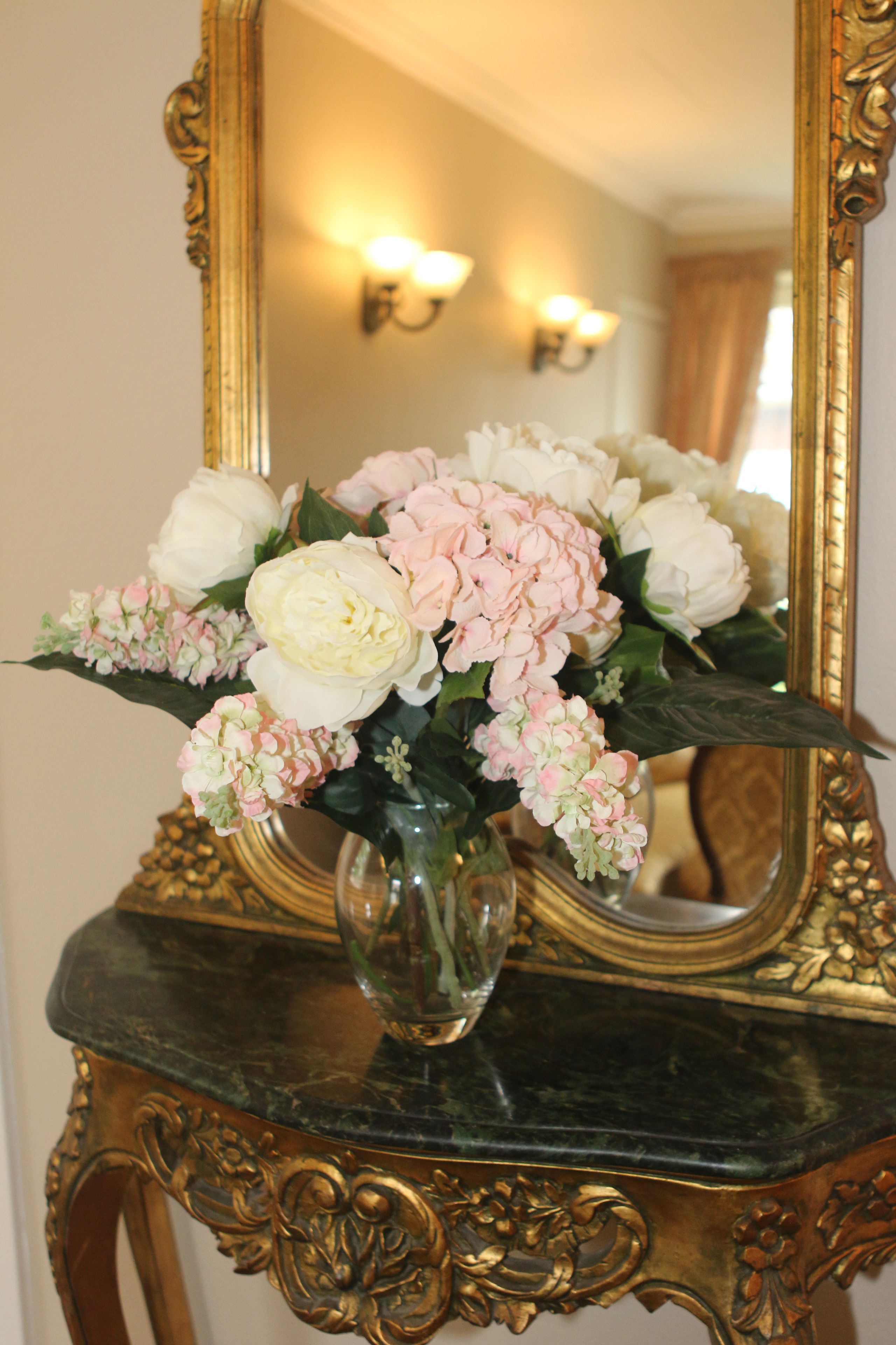 A mirror with a gold frame, a vase with flowers, and a marble table.