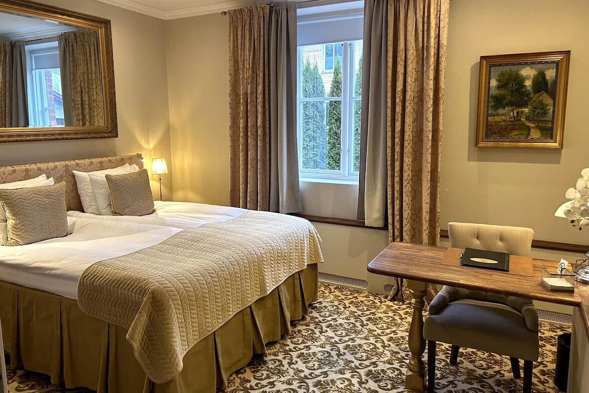 Une chambre d’hôtel avec un grand lit, un bureau avec une chaise, un tableau au mur et une fenêtre avec des rideaux.