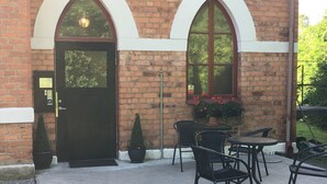 A brick building with a black door and two arched windows. There are outdoor tables and chairs, and a potted plant near the windows. - Sockerslottet hotell (Kristinehamn)