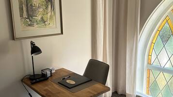 A hotel room with a wooden desk, a chair, a lamp, a trash can, a framed picture on the wall, and a window with a stained glass view.