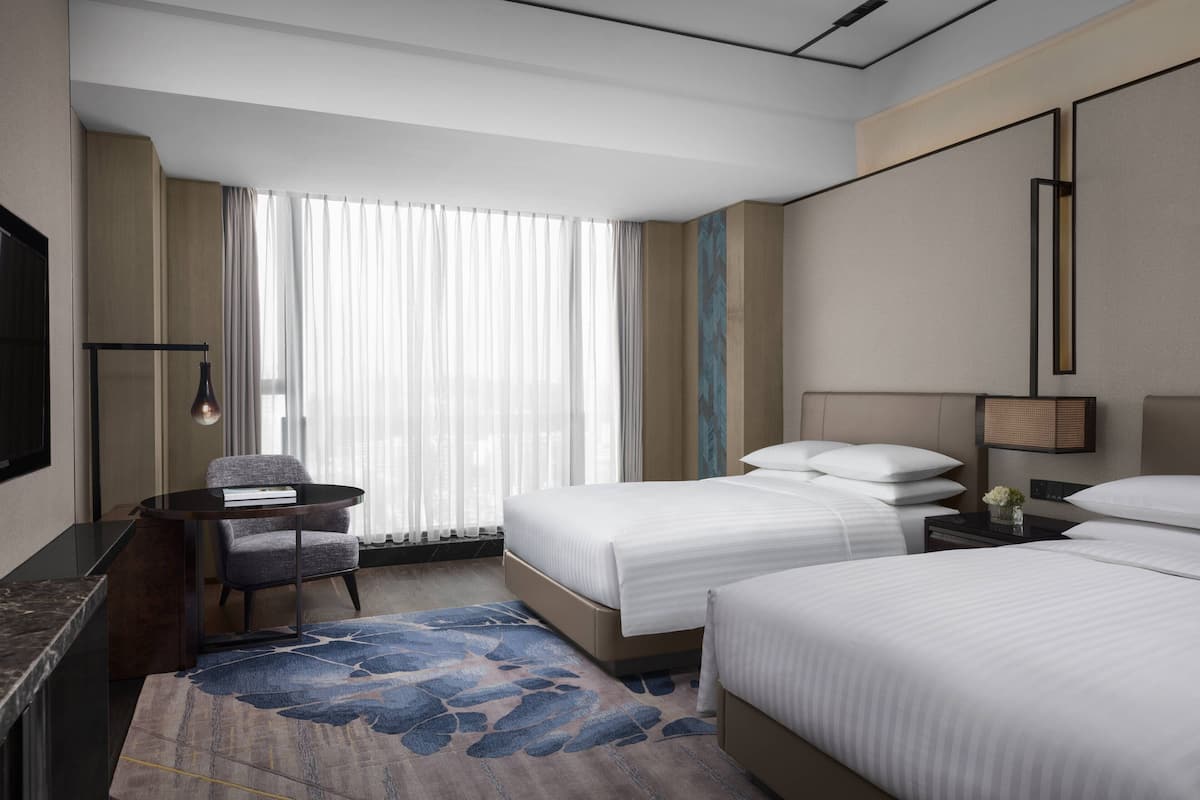 Ein Hotelzimmer mit zwei Betten, einem Schreibtisch, einem Stuhl, einem Fernseher und einem großen Fenster mit Vorhängen.
