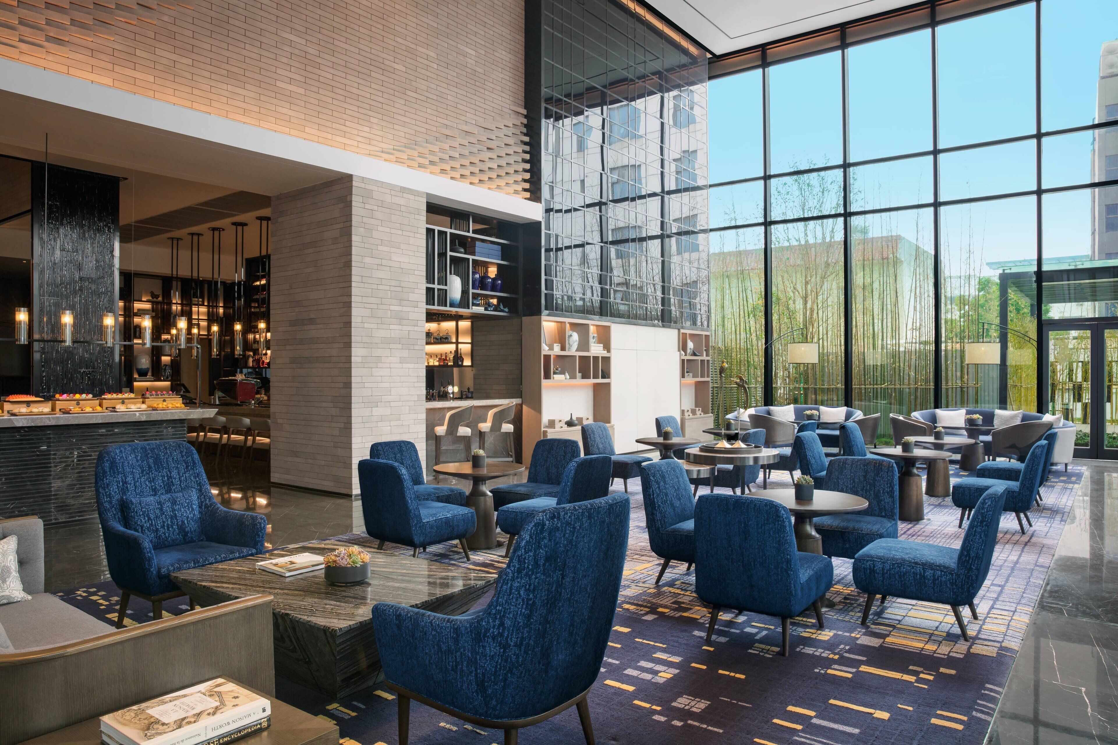 A modern lobby with blue upholstered chairs, a bar area, and large windows offering a view of the outside.