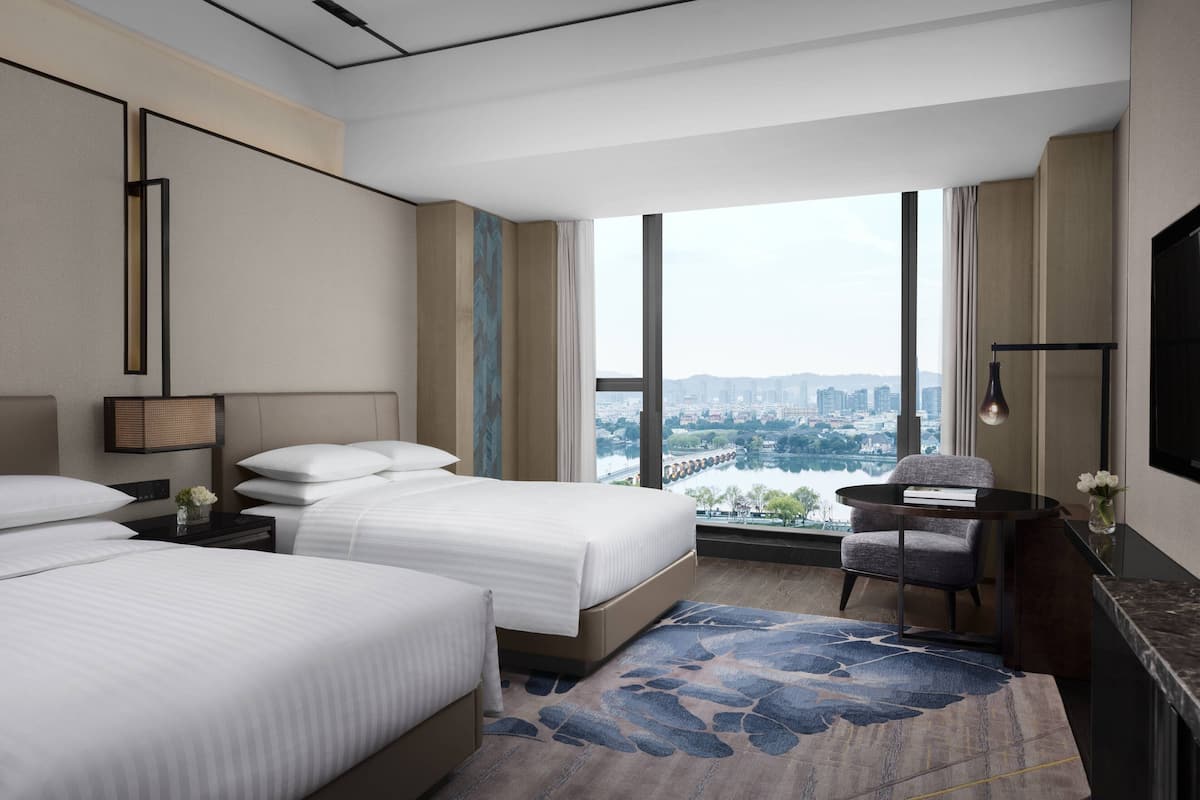 Ein Hotelzimmer mit zwei Betten, einem Schreibtisch, einem Sessel, einem Fernseher und einem großen Fenster mit Blick auf die Stadt.