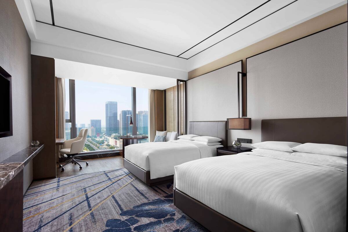 Ein Hotelzimmer mit zwei Betten, einem Schreibtisch, einem Sessel und einem großen Fenster mit Blick auf die Stadt.