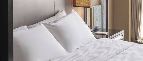 A neatly made bed with white linens and pillows, a modern lamp on a bedside table, and a window with curtains.