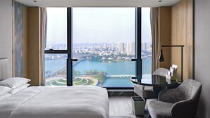 Une chambre d’hôtel moderne dotée d’un grand lit, d’un coin salon avec une table et offrant une vue sur le paysage urbain et un plan d’eau grâce à de grandes fenêtres du sol au plafond.