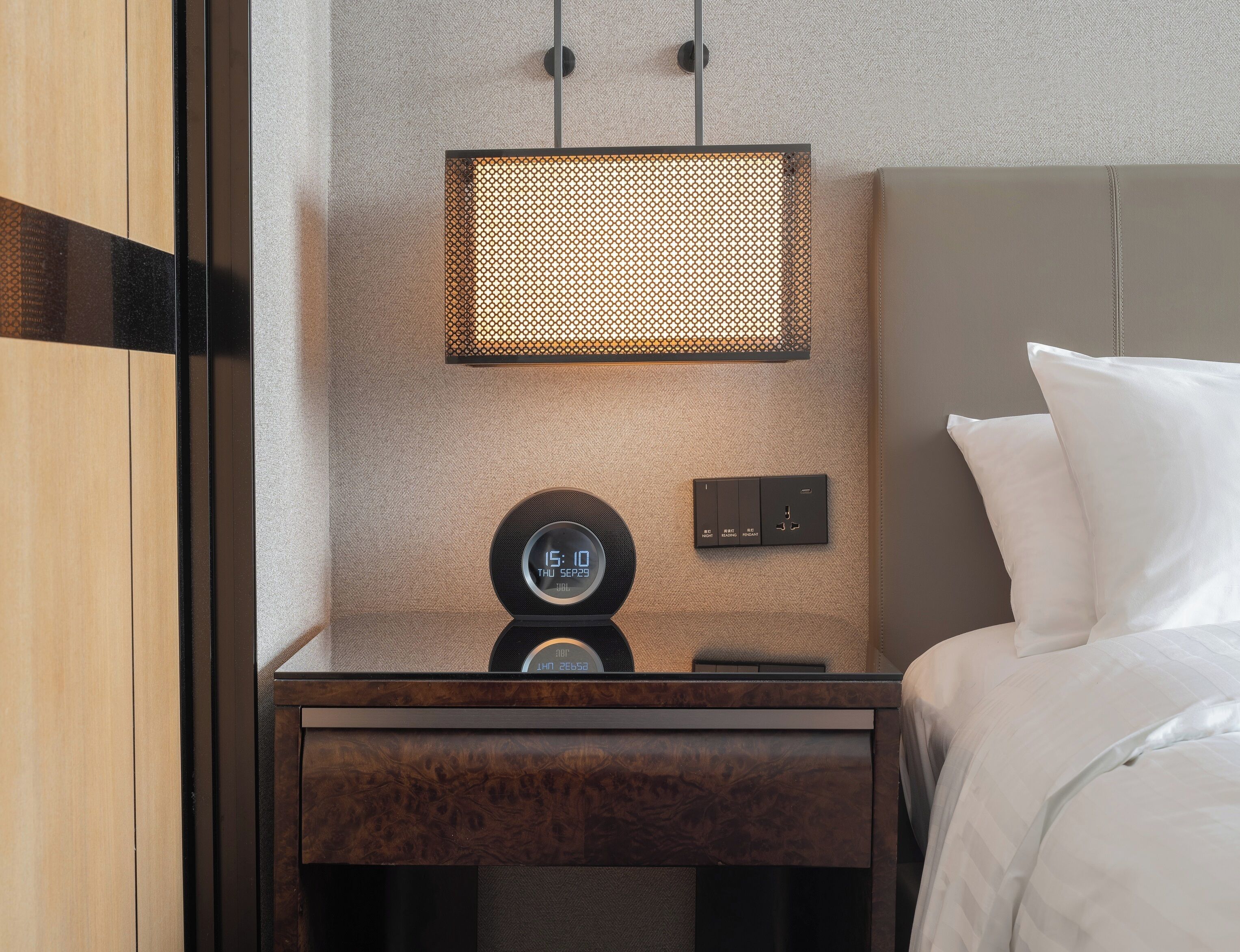 Una cama bien tendida con sábanas blancas, una mesita de noche con un reloj y un enchufe, y una lámpara fijada a la pared.