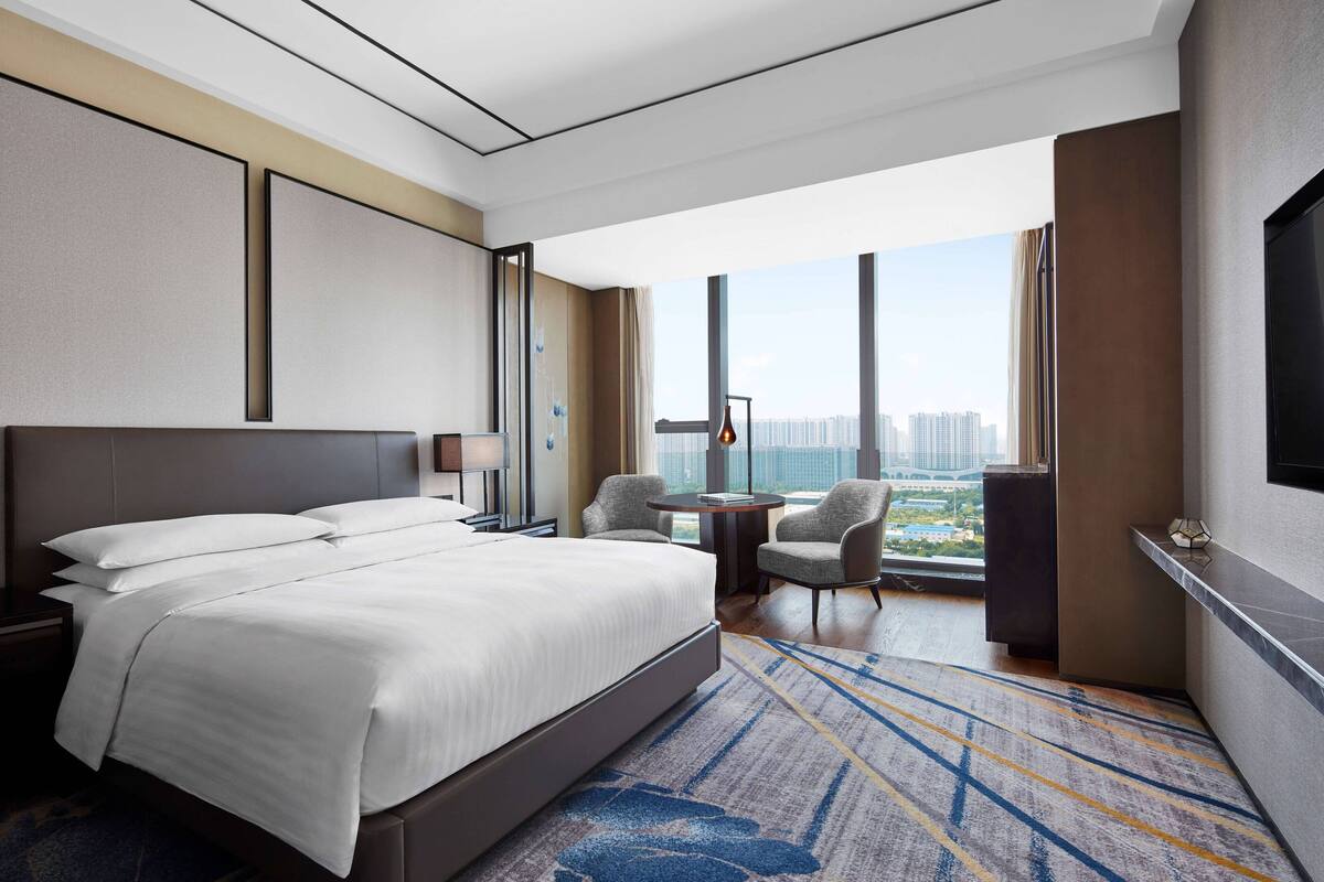 Ein modernes Hotelzimmer mit einem großen Bett, einem Schreibtisch mit Stuhl, einem Fernseher und Blick auf die Stadt.