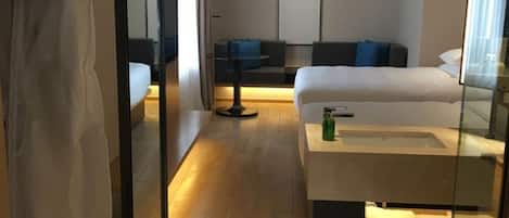 Une chambre d’hôtel moderne dotée d’un grand lit, d’une table de chevet, d’un miroir et d’un coin salon avec un canapé et une petite table.