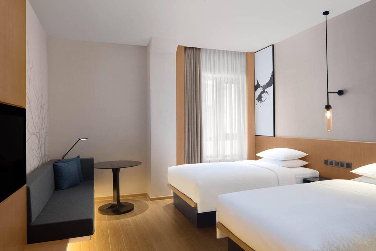 A hotel room with a sofa, two beds, a small table, and a wall-mounted light fixture.