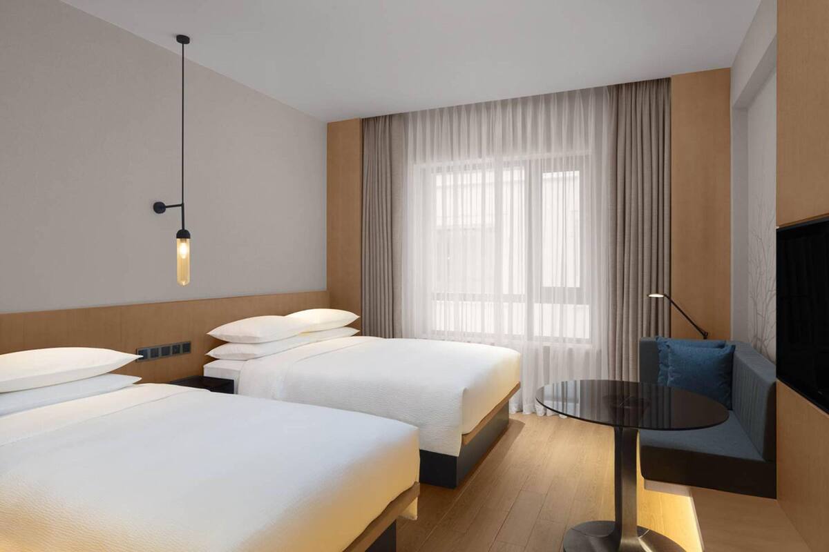 A hotel room with two beds, a round table, a sofa, a TV, and a large window with sheer curtains.