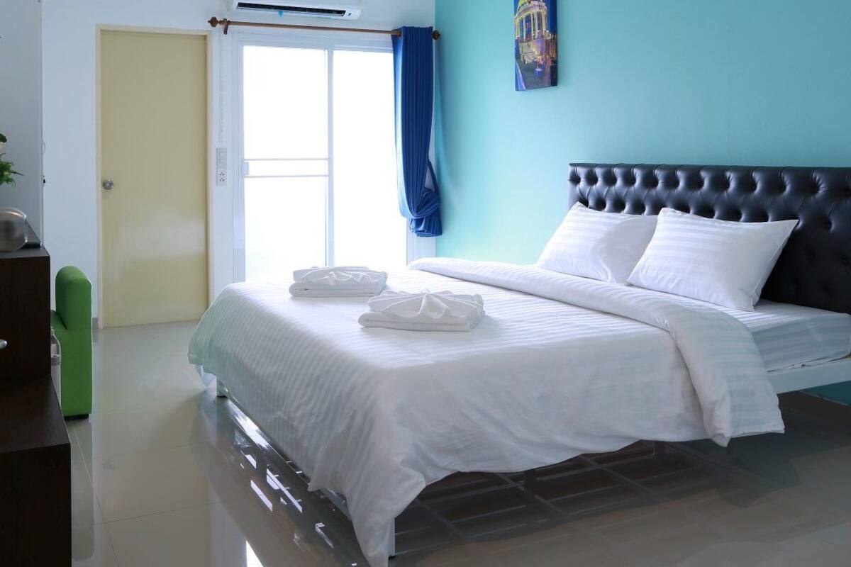 A hotel room with a bed, a ceiling fan, an air conditioning unit, a painting on the wall, and a window with curtains.