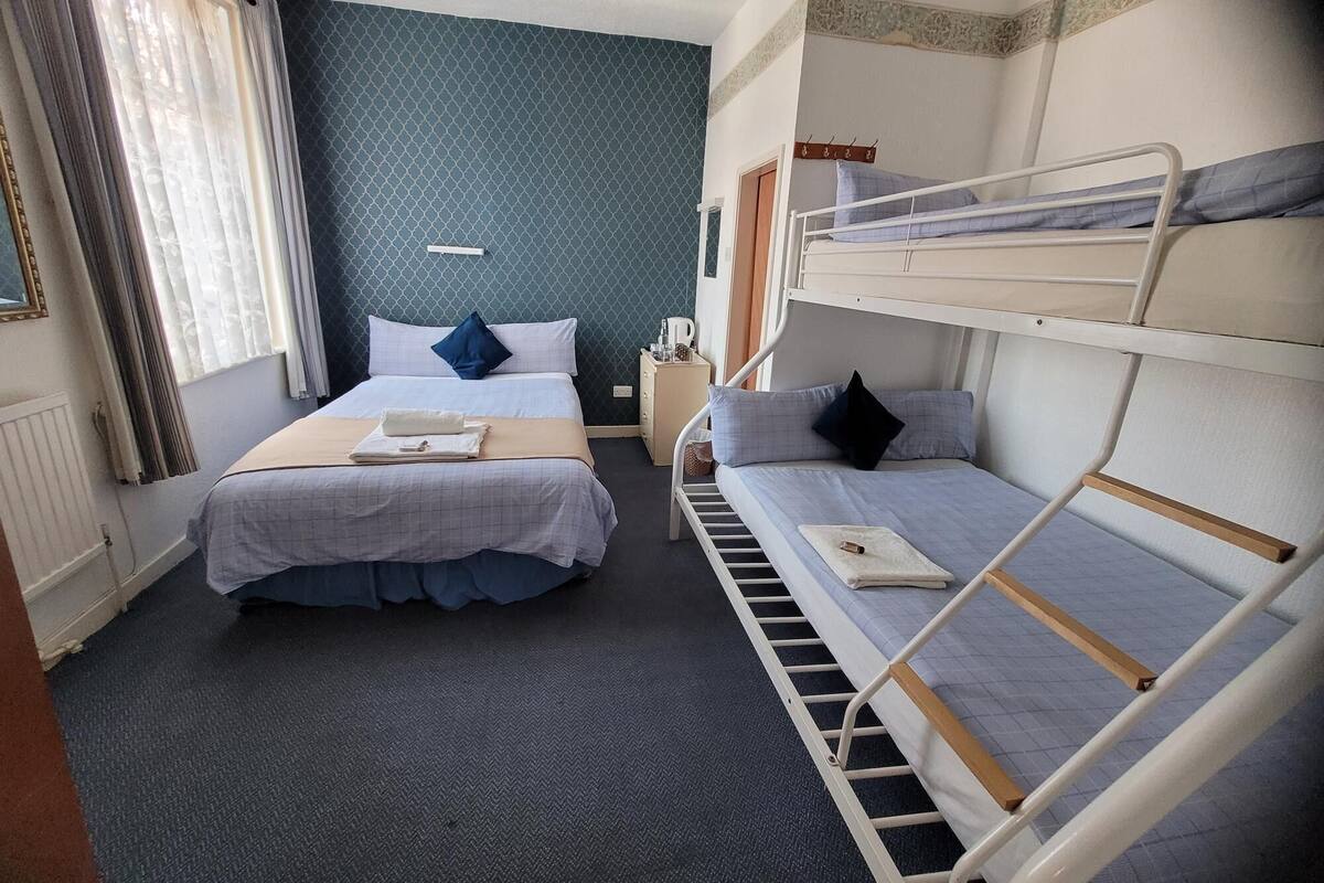 Une chambre d’hôtel avec deux lits, dont un à baldaquin et un autre superposé. Il y a une petite table de chevet avec une lampe et une tasse. La chambre a un mur bleu foncé et une fenêtre avec des rideaux.