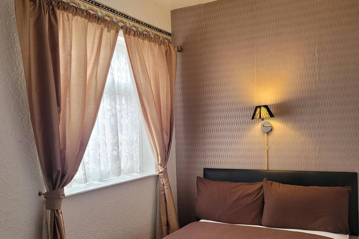 Une chambre d’hôtel avec un lit, des rideaux, une lampe fixée au mur et une fenêtre.