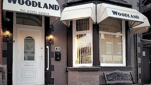 A building with a sign reading "WOODLAND" and a telephone number. There is a bench outside and a door with a number "50" above it. - The Woodland Hotel (Blackpool)