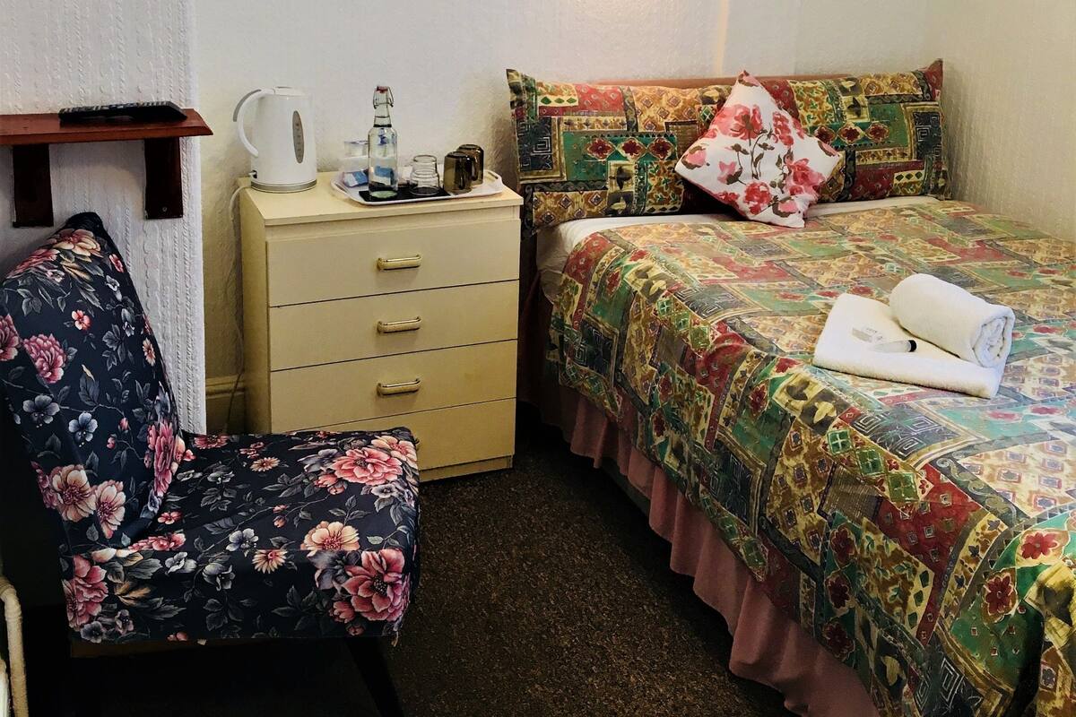 Une petite pièce agréable, dotée d’un lit, d’une chaise, d’une table de chevet et d’une lampe fixée au mur.