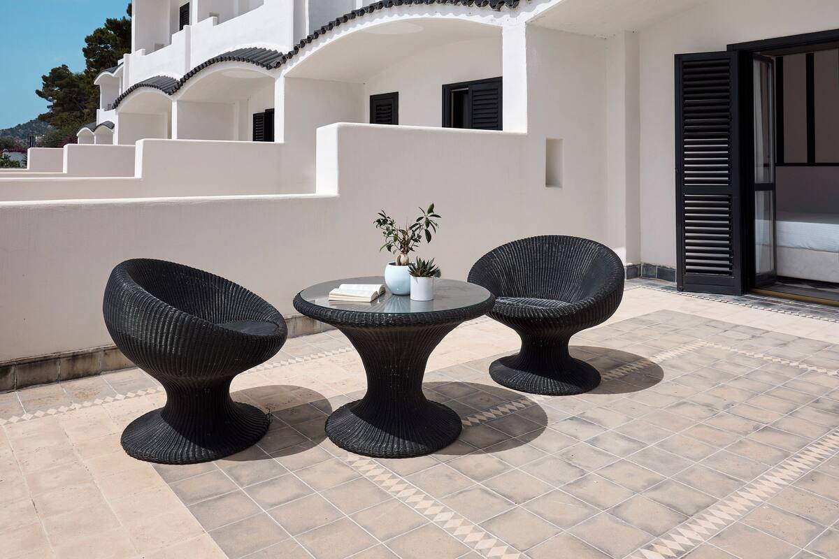 Ein modernes Gebäude mit weißer Fassade, großen Fenstern und einer gefliesten Terrasse mit zwei Korbstühlen und einem Tisch.
