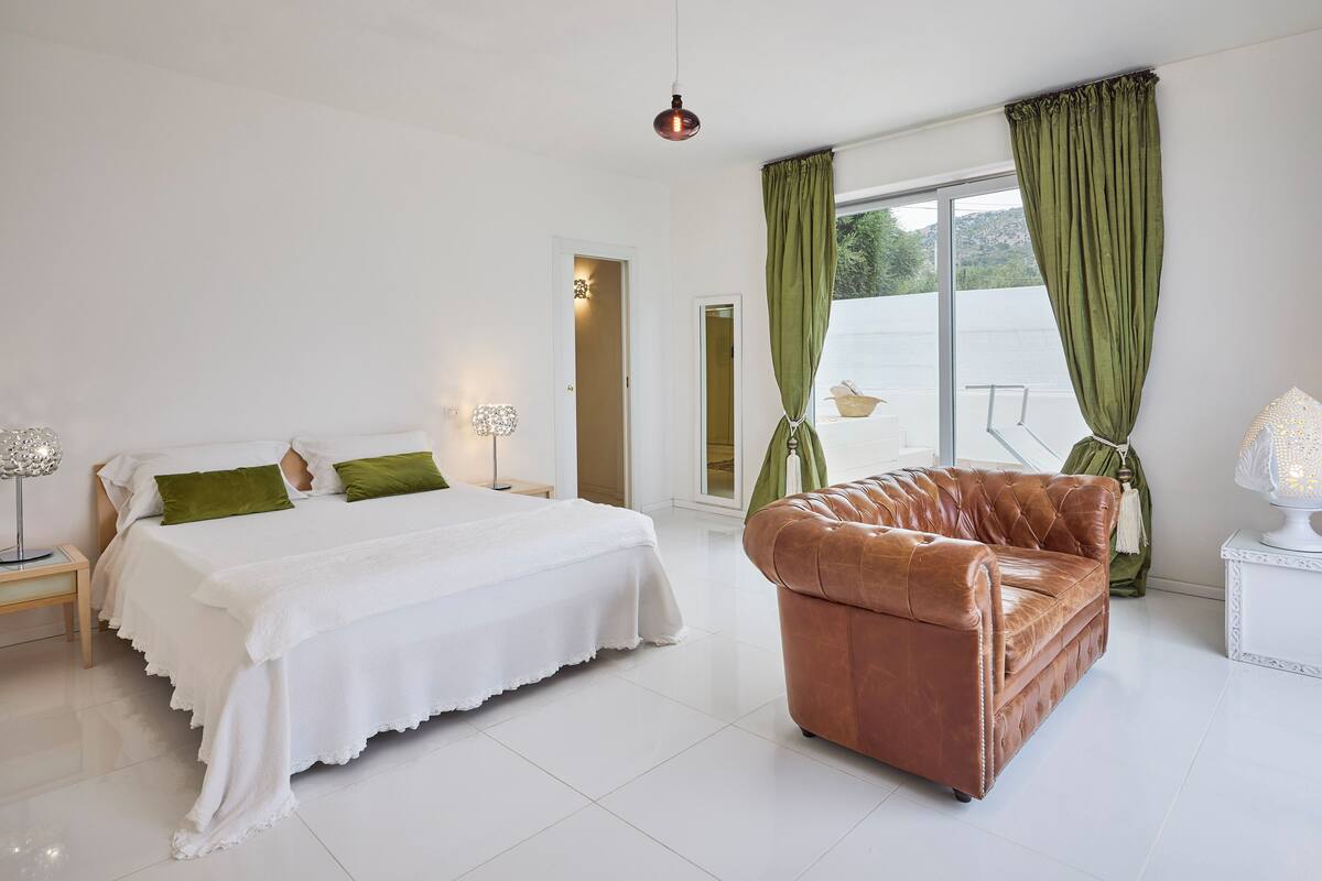 Ein Schlafzimmer mit einem Bett, einem braunen Ledersofa, einem Fenster mit grünen Vorhängen und einer weißen Lampe.