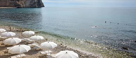 Una playa con sombrillas blancas y sillas de descanso, una costa rocosa y un acantilado al fondo.