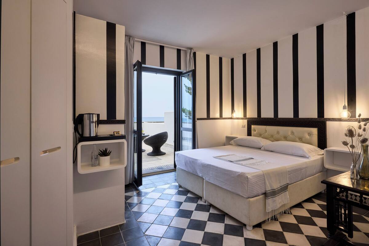 Ein Schlafzimmer mit Schachbrettmuster-Boden, einem Bett mit weißem Kopfteil, einem Nachttisch mit Lampe und Blick durch eine offene Tür.