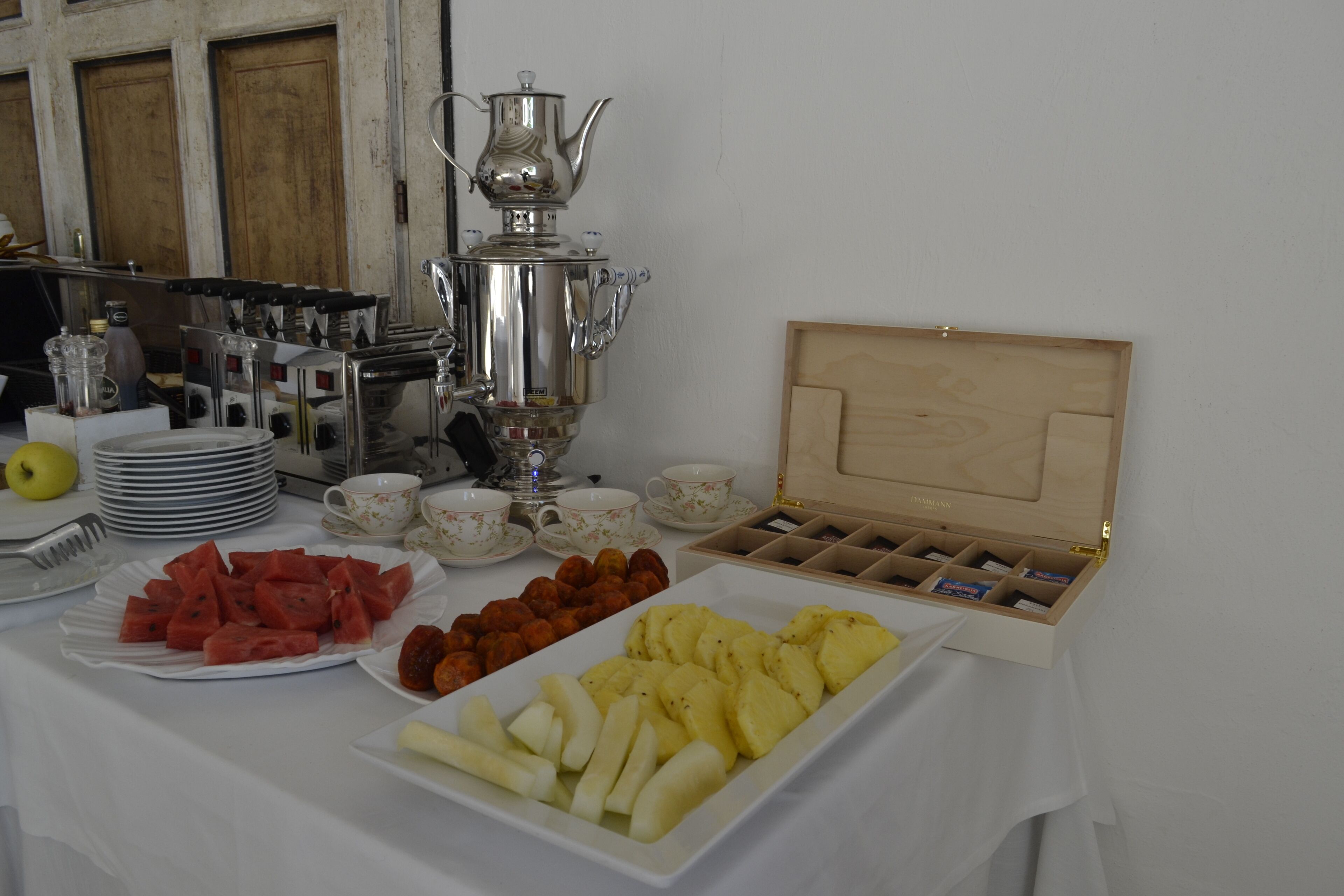 A buffet table with plates of watermelon, bananas, and other fruits, a silver teapot, and a wooden box with compartments.