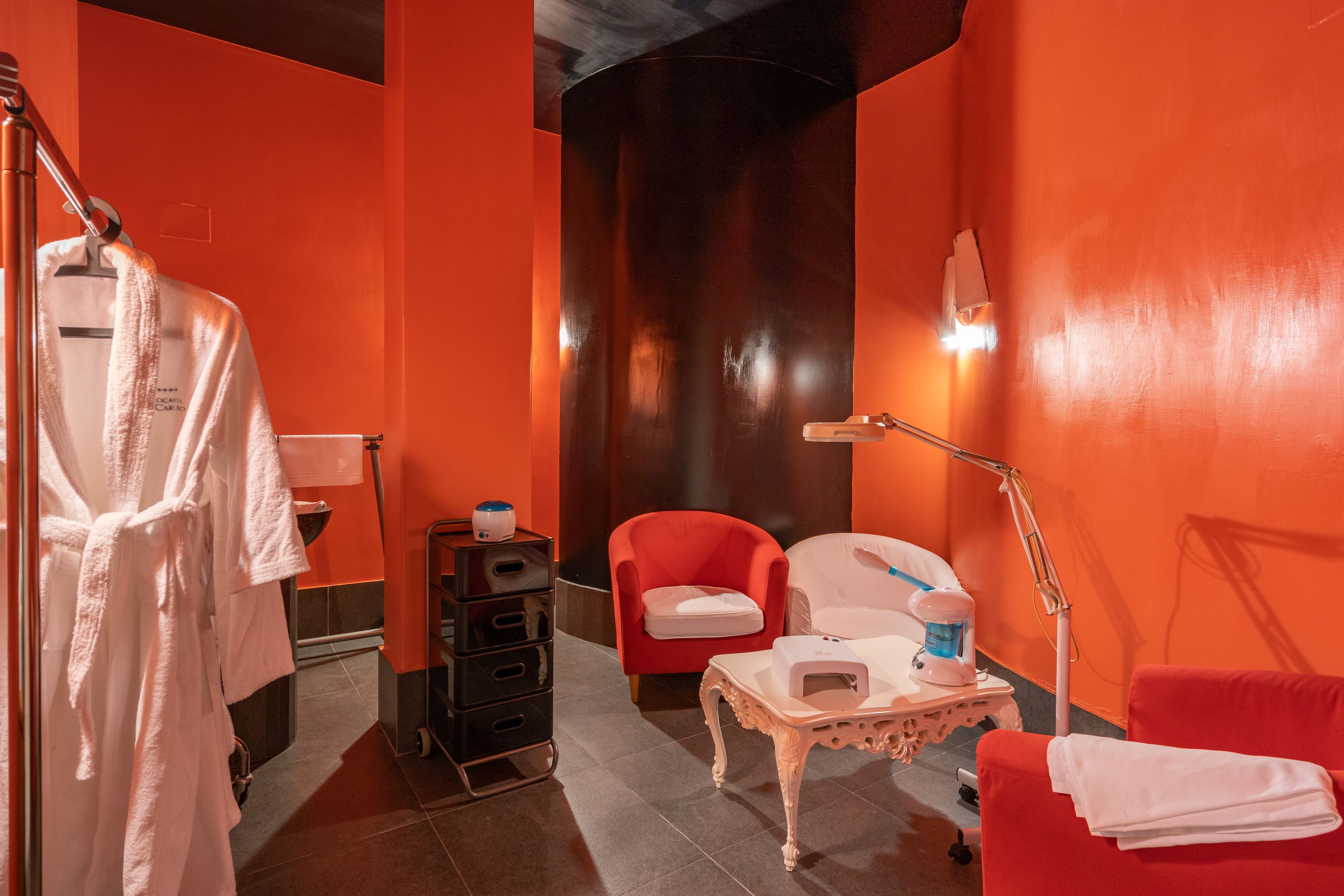 Une pièce aux couleurs rouge et noir, avec un fauteuil rouge, une table blanche surmontée d’une lampe, et une robe blanche suspendue à un porte-manteau.
