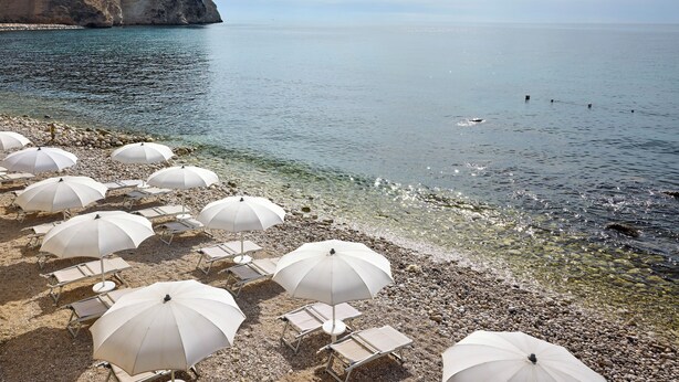 A beach with white umbrellas and lounge chairs, a rocky shoreline, and a cliff in the background. - La Locanda del Carrubo (Mattinata)