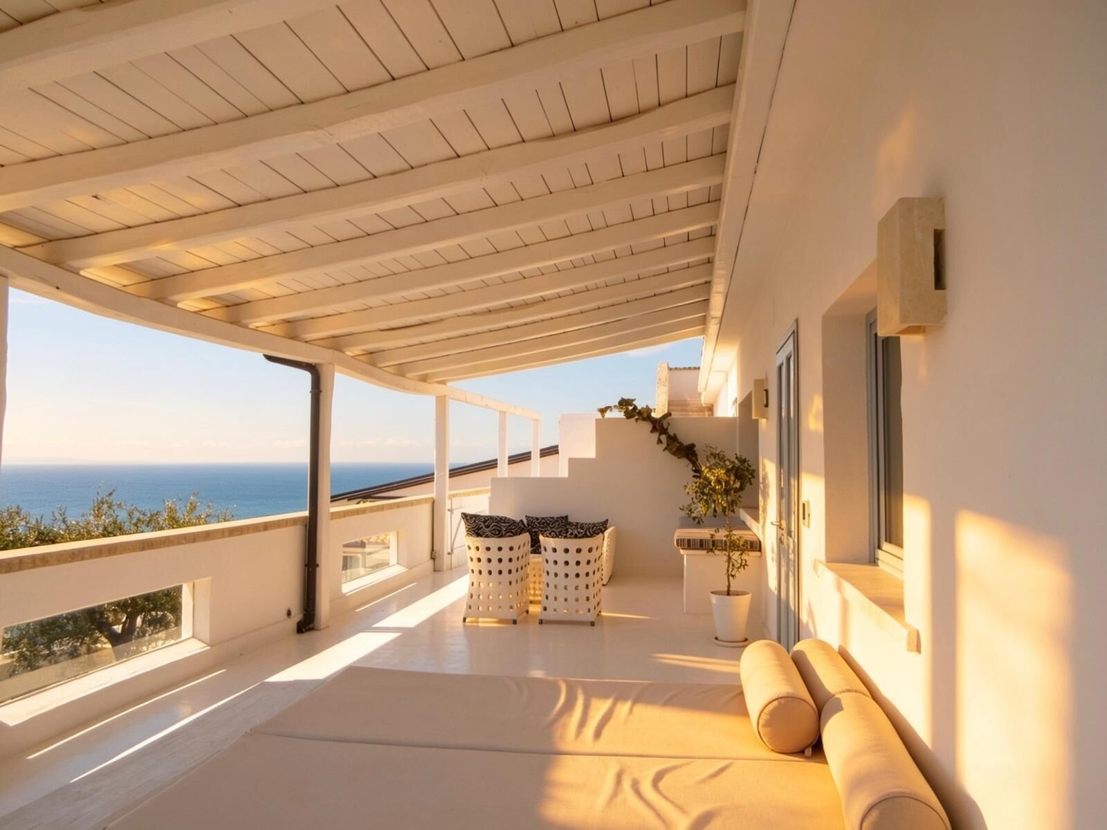 A sunlit balcony with a view of the sea, a seating area with cushions, and a potted plant.