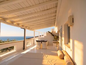 Un balcon ensoleillé avec vue sur la mer, un coin salon avec des coussins et une plante en pot.
