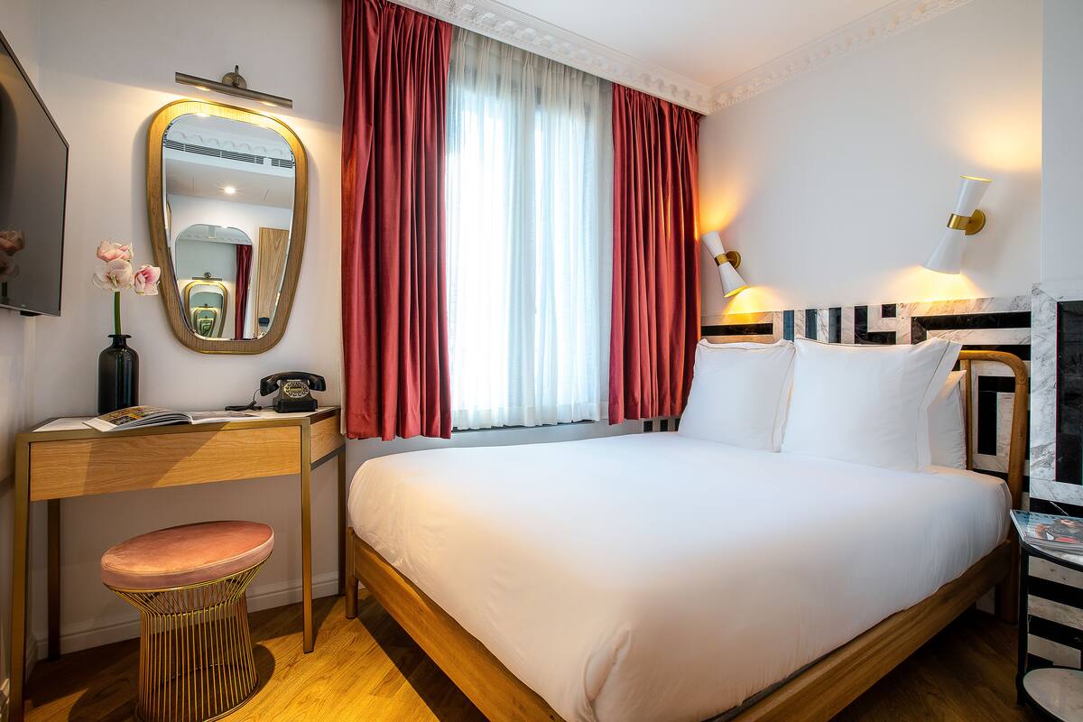 Habitación de hotel con cama, escritorio con teléfono, espejo y ventana con cortinas.