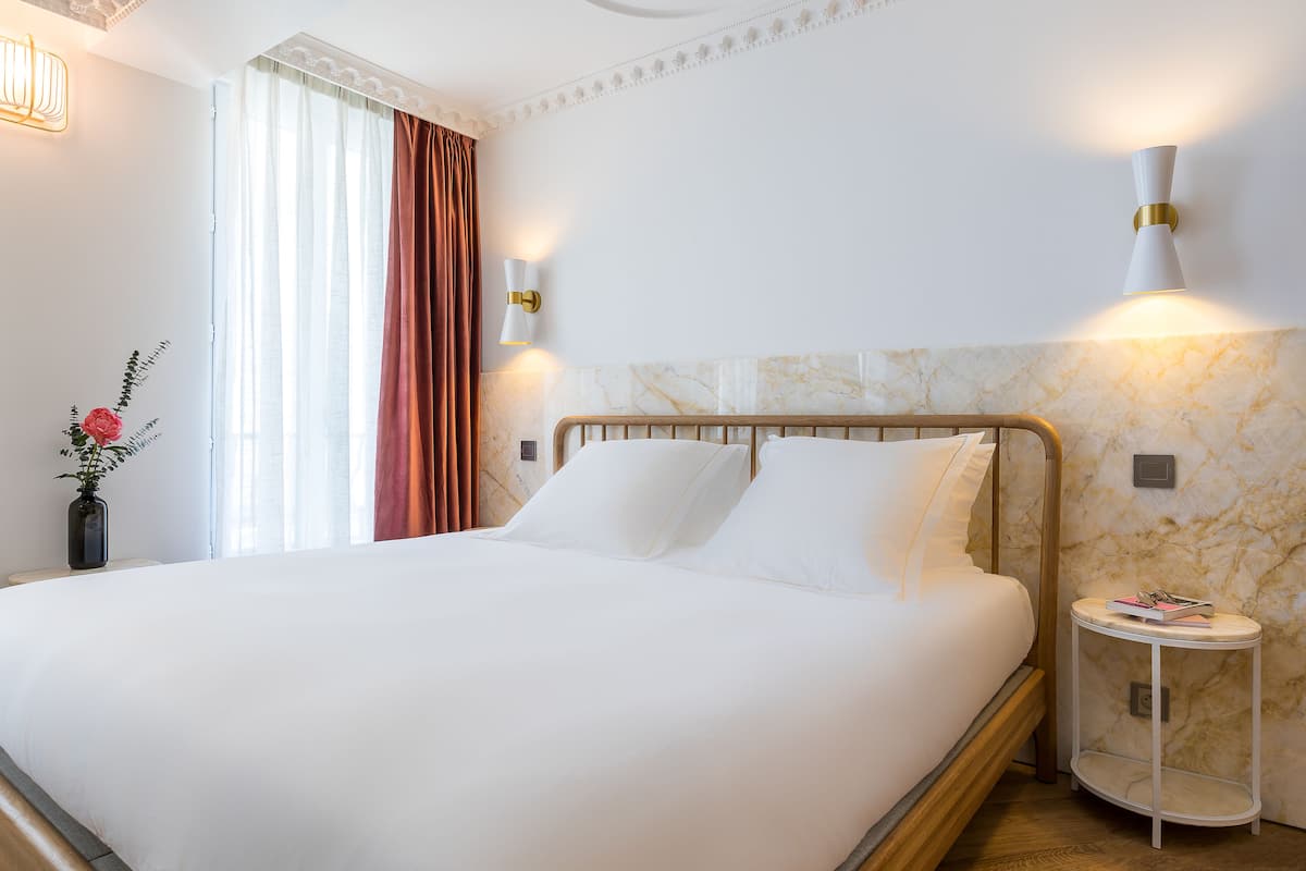 Una cama bien hecha con ropa blanca, una mesita de noche con un libro y un florero, y lámparas fijadas a la pared.