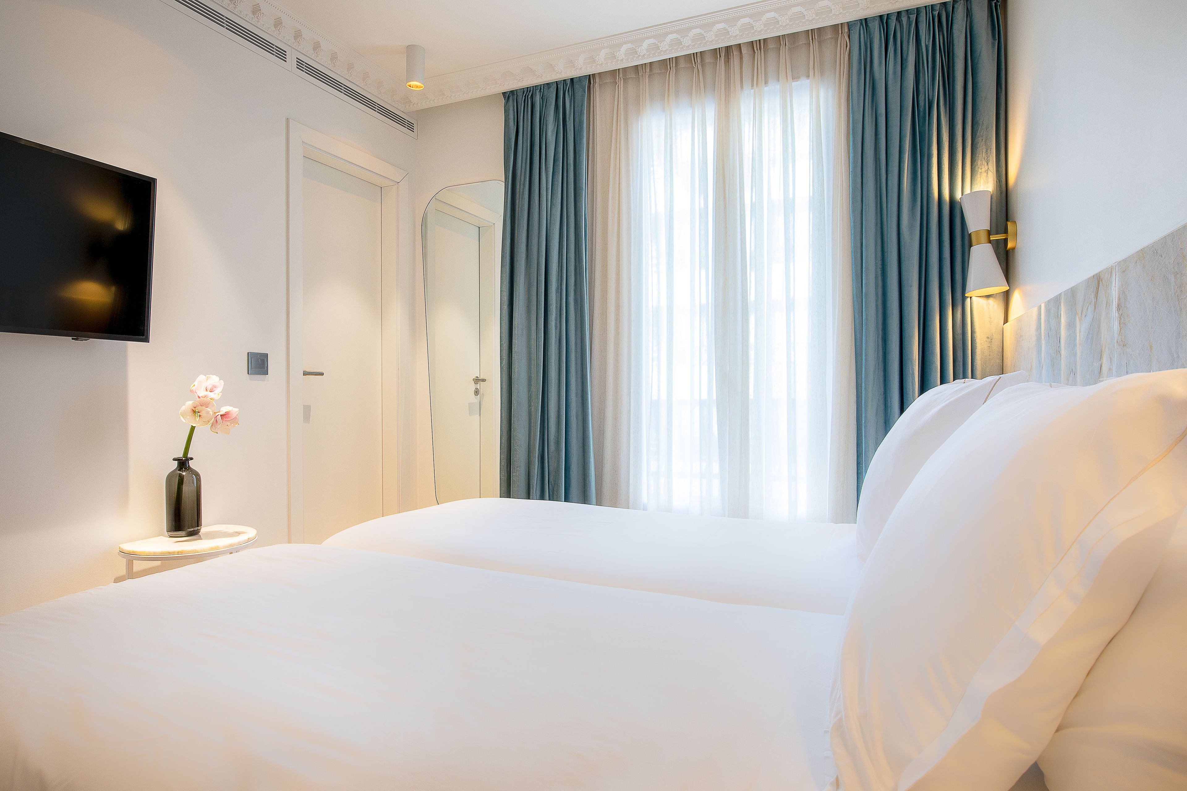 A hotel room with a large bed, a TV on the wall, and a vase with flowers on a bedside table.