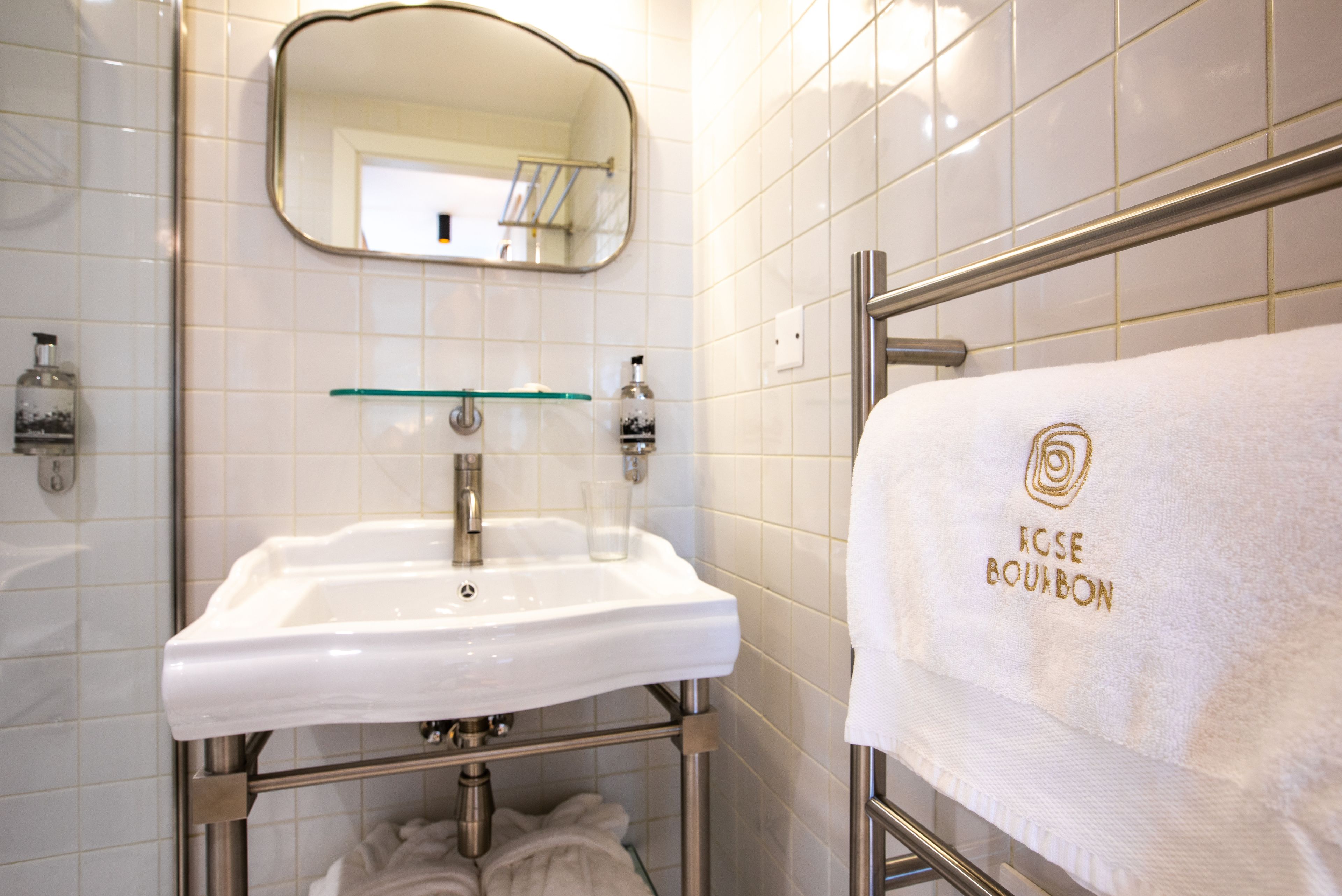 Un bagno con un lavandino bianco, uno scaffale in vetro, un portasacchiuni con un asciugamano e uno specchio.