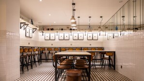 Um restaurante com chão quadriculado, cadeiras de madeira, uma mesa central e quadros nas paredes.
