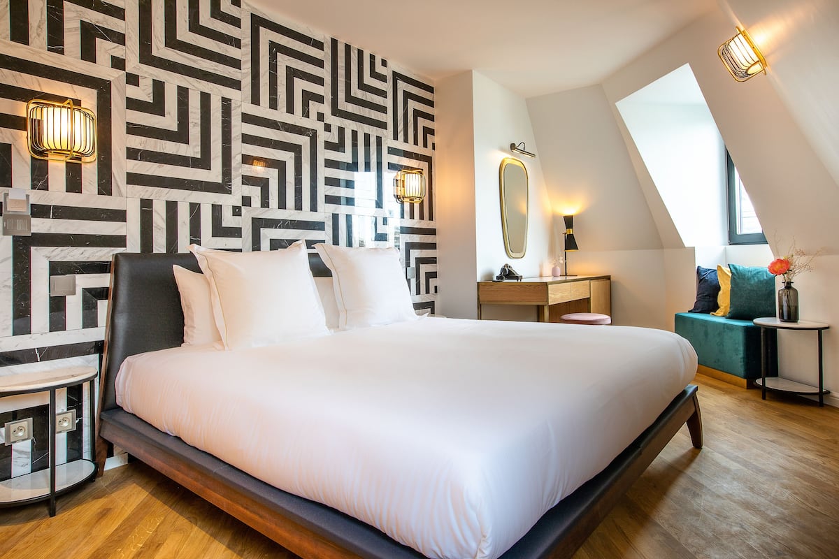 Una habitación de hotel moderna con una cama grande, un muro con diseño geométrico, una tocador con espejo y un tragaluz.