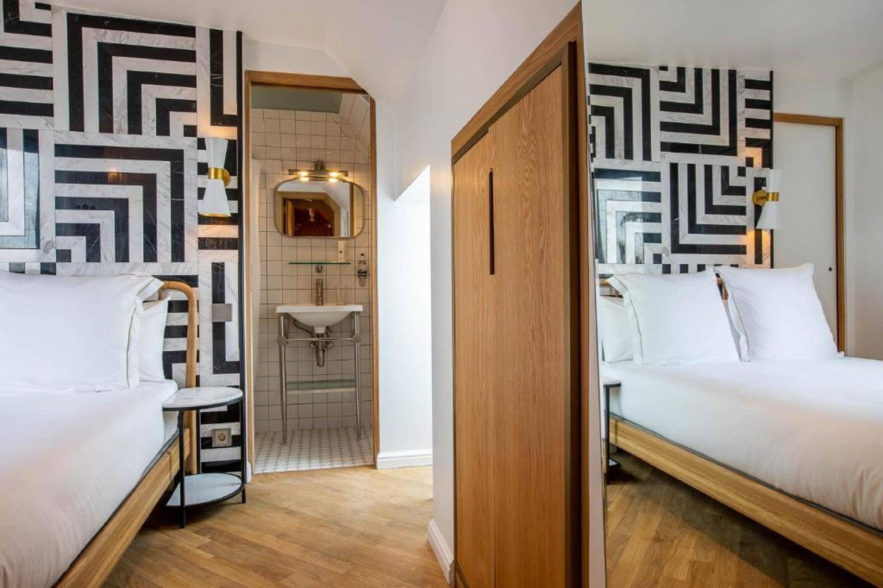 A hotel room with two beds, a wooden floor, a bathroom with a sink and mirror, and a geometric patterned wall.