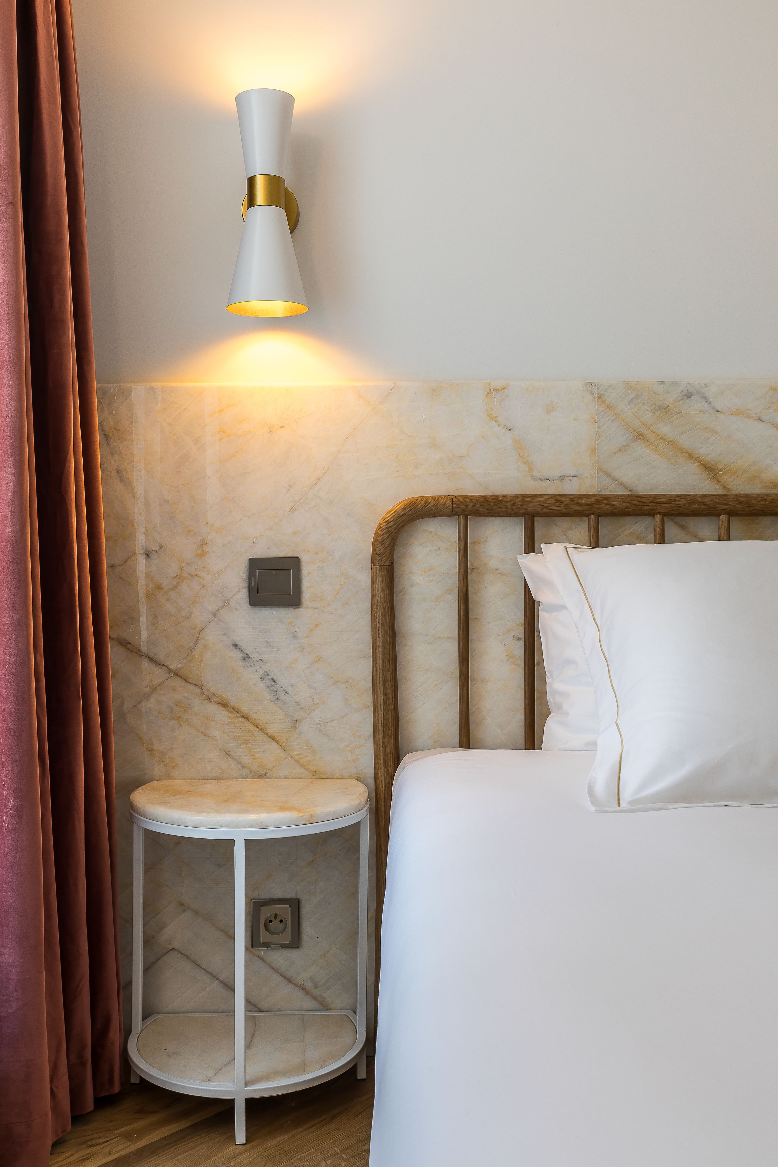 A hotel room with a bed, a bedside table, a wall-mounted lamp, and a marble-patterned wall.