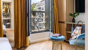 Quarto moderno com uma janela grande, varanda, cama, poltrona azul e uma pequena mesa com livros.