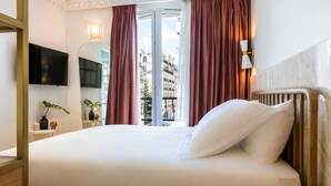 Quarto de hotel com uma cama grande, uma televisão fixada na parede, uma janela com cortinas e vista para edifícios.