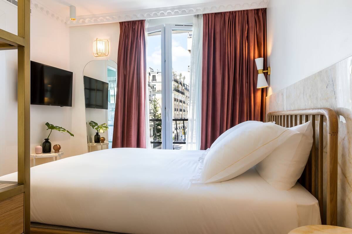 Habitación de hotel con una cama grande, televisión montada en la pared, ventana con cortinas y vista a edificios.