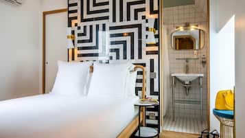 A hotel room with a bed, a bedside table, a mirror, a sink, and a shower.