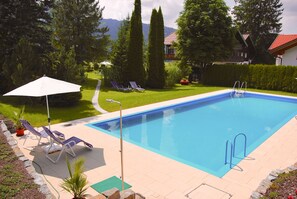 Una piscina rectangular con escalera, rodeada por un deck pavimentado. Hay dos sillas reclinables y una sombrilla blanca.