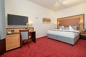 Habitación de hotel con cama, escritorio, silla, televisor y piso cubierto de alfombra roja.