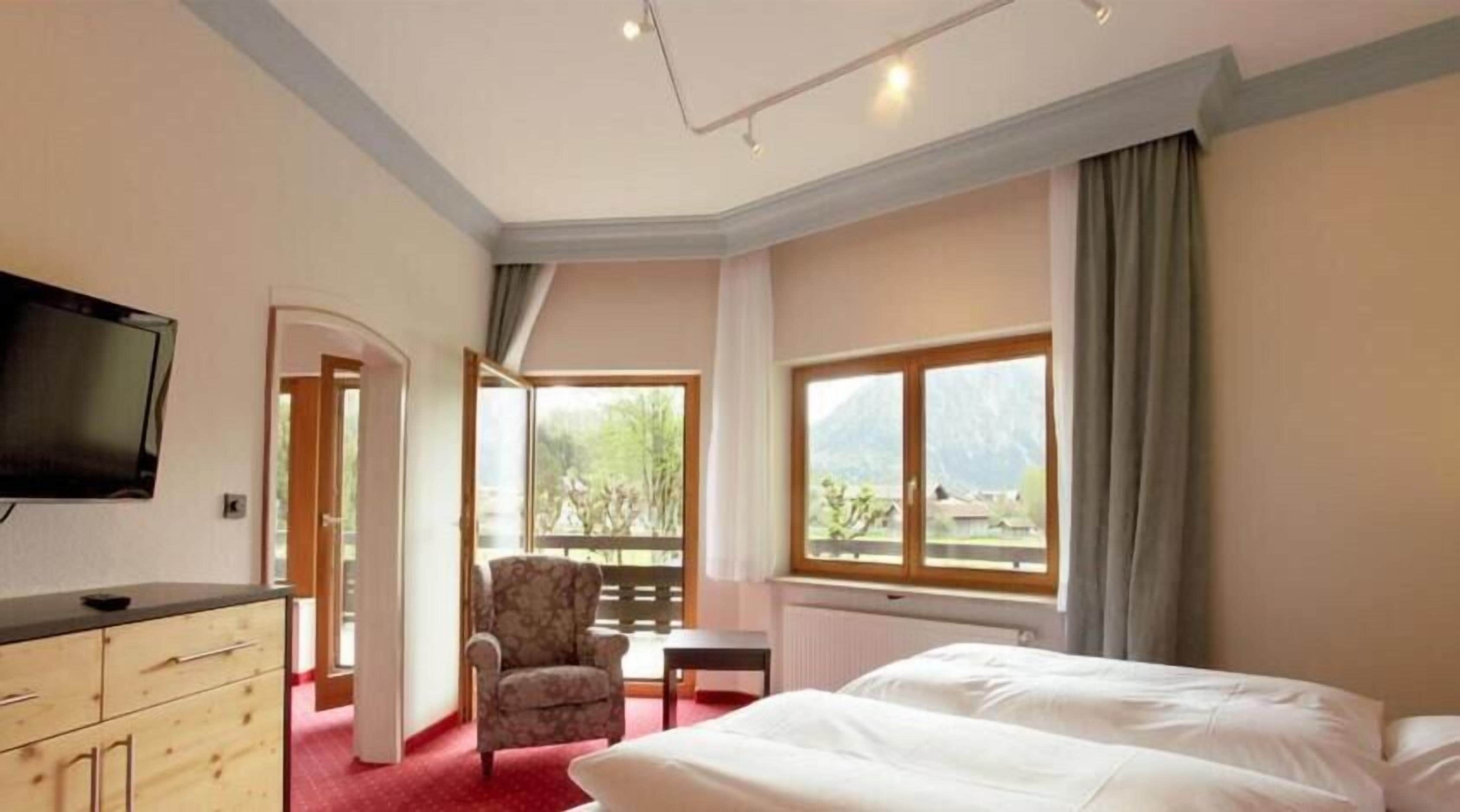A hotel room with a bed, a chair, a small table, a TV, and a window with a mountain view.