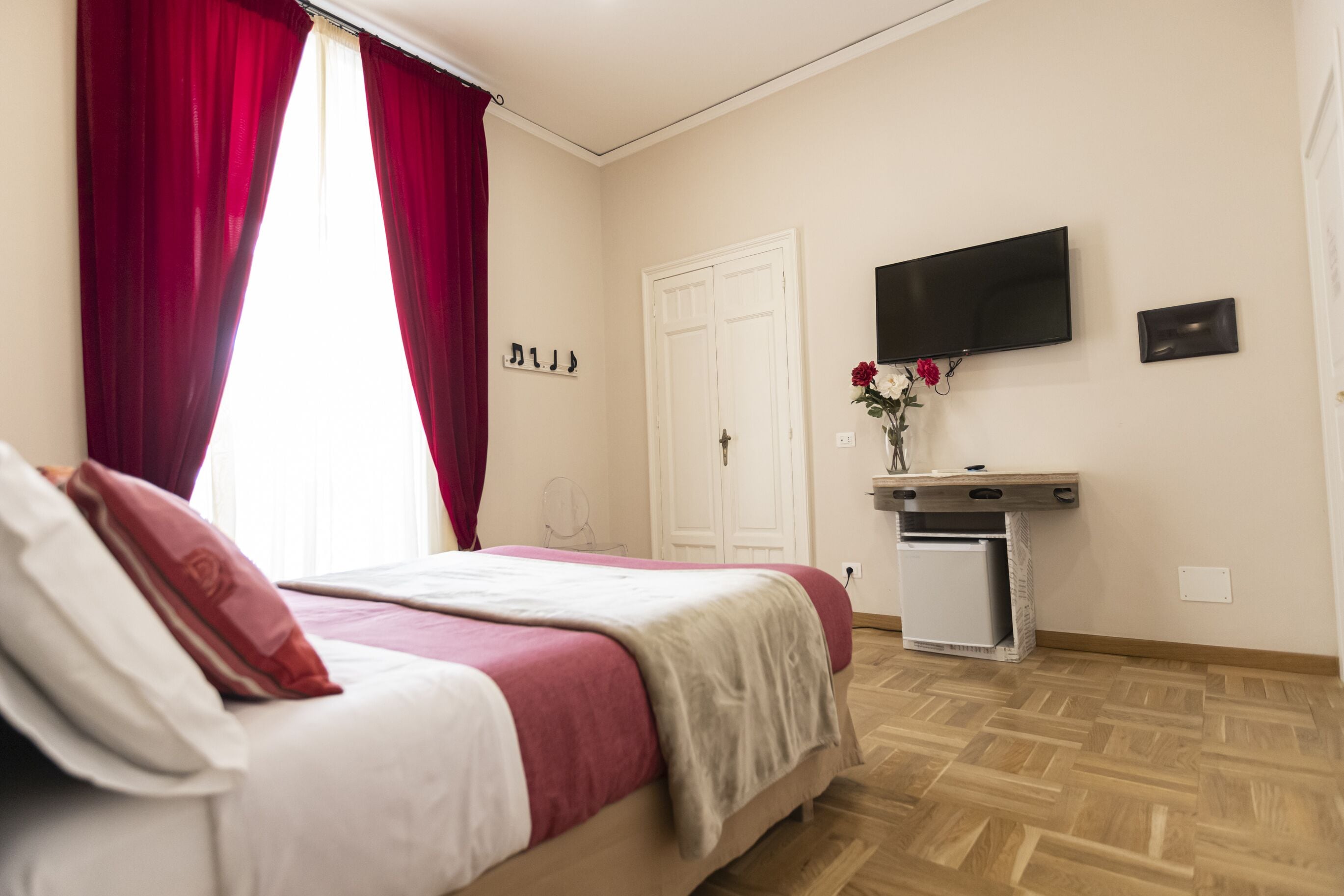 Un dormitorio con cama, televisor montado en la pared, un escritorio pequeño y una ventana con cortinas rojas.
