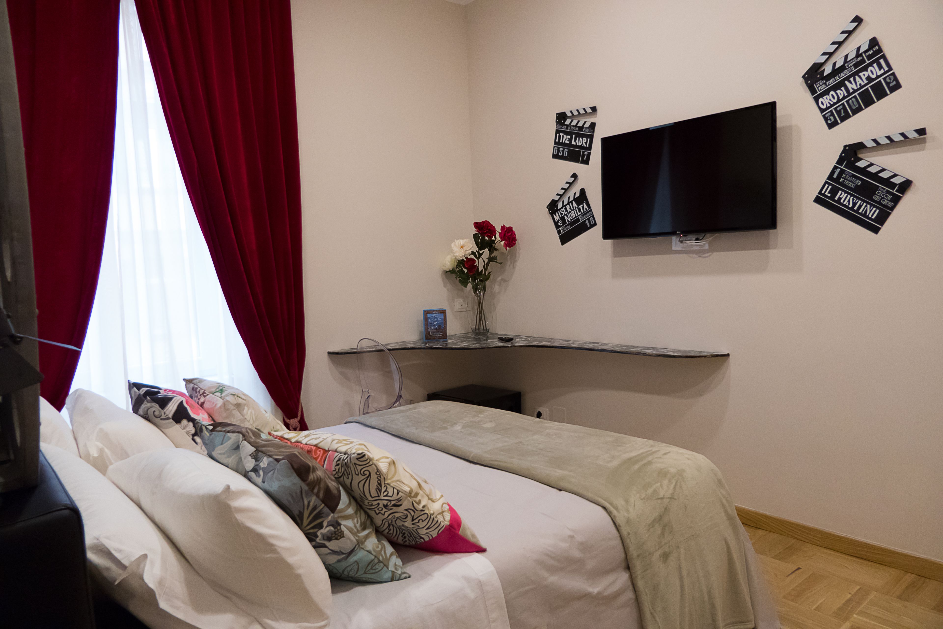 Quarto de hotel com uma cama, televisão na parede e escrivaninha com um vaso de flores.