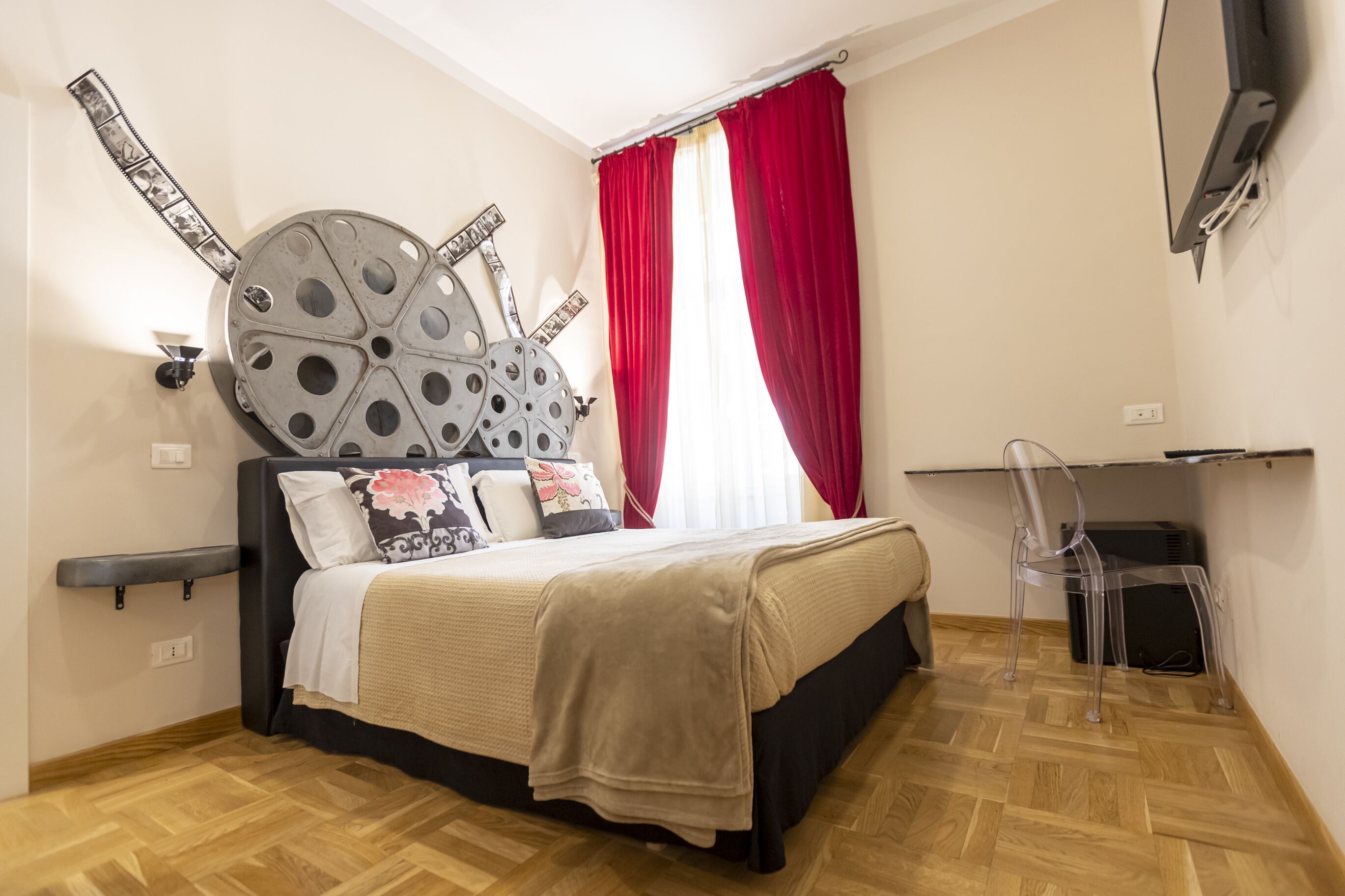 Un dormitorio con cama, cabecera con diseños circulares, cortina roja, una silla transparente y un televisor montado en la pared.