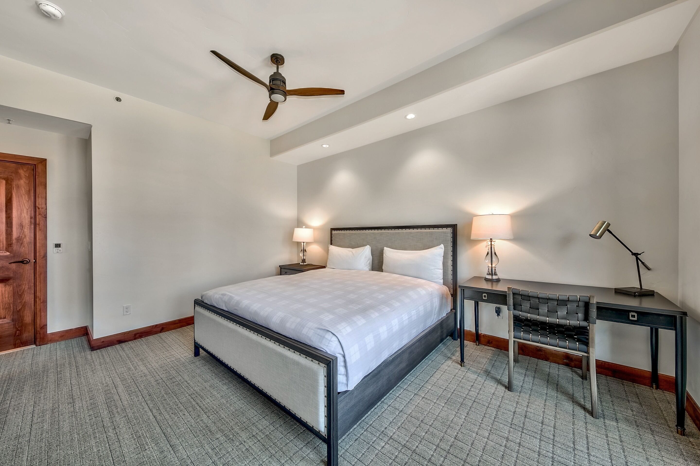 A bedroom with a bed, bedside tables, a desk, a chair, a ceiling fan, and a door.
